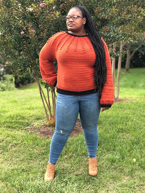 A woman stands in a field with trees, wearing a bold orange crocheted sweater with a bobble design on the yoke.