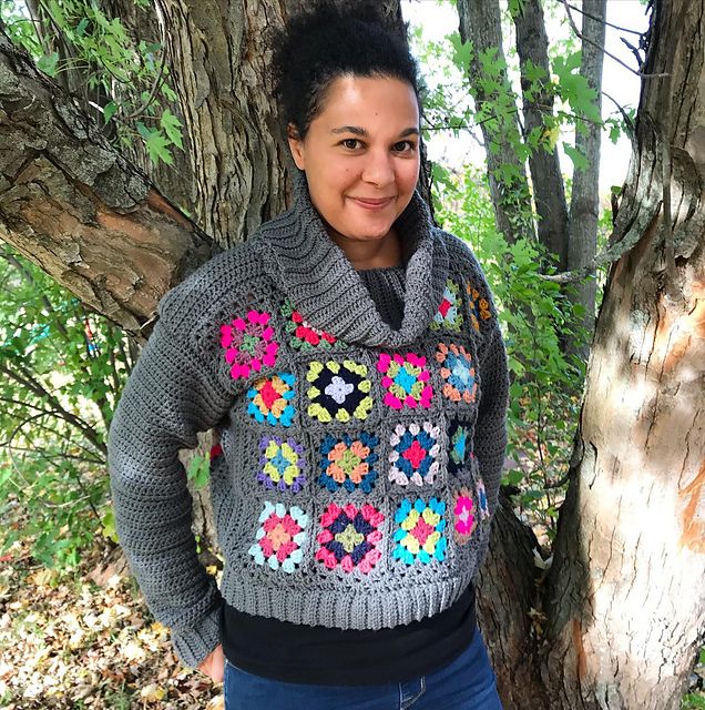 A woman wearing a crocheted cowl-neck sweater in grey with bright granny squares stands in front of a tree, smiling at the camera. 