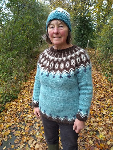 A woman in an aqua sweater with chocolate brown and white colorwork stands on a forest path covered in yellow leaves.