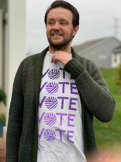 A man in a rich green cardigan and a VOTE t-shirt smiles off-camera.