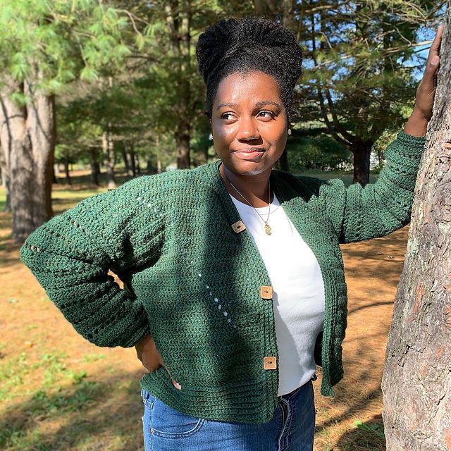 A woman leans against a tree, smiling and wearing a green crocheted cardigan.