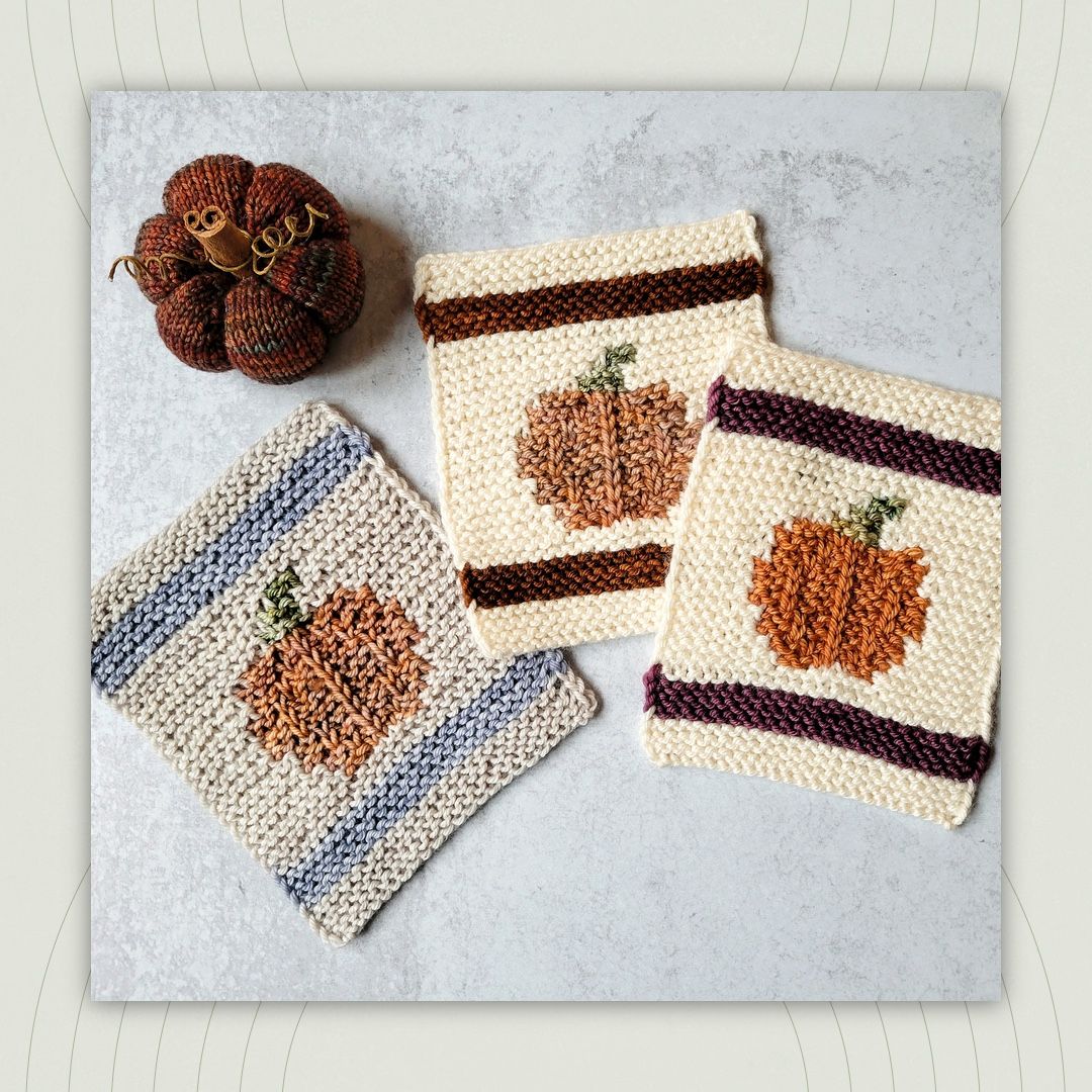 three handknit garter stitch coasters with bold stripe borders and textured pumpkin designs in the middle, arranged in a flat-lay. a rust-colored handknit pumpkin with a cinnamon stick stump is on the table just above them.  
