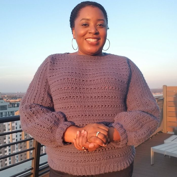 A smiling woman stands in glowing light on a rooftop patio, her hands clasped in front of her, wearing a purple crocheted sweater with puffed sleeves, accessorized with delicate rings and hoop earrings.