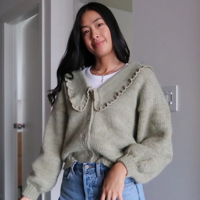 A woman stands, smiling at the camera, her head tilted and long hair flowing over her shoulder. She is wearing a sage green cardigan with puffed sleeves and a ruffled v-neck.