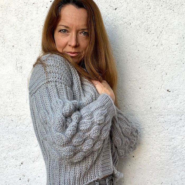 A woman stands with her body facing to the side and head confidently looking at the camera. Her grey cardigan sweater is handknit and richly textured, with puffed sleeves.