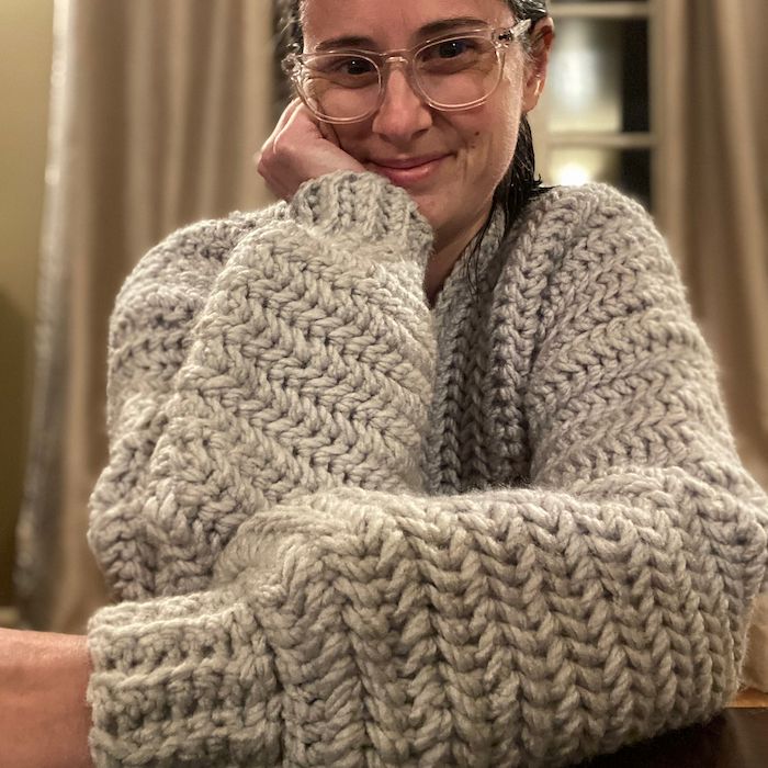 A close-up photo of a woman sitting at a table, one arm with elbow propped on the table and hand on her chin, the other arm resting on the table. She's smiling and wearing a bulky crocheted sweater with a rich texture, oatmeal grey color, and puffed sleeves.