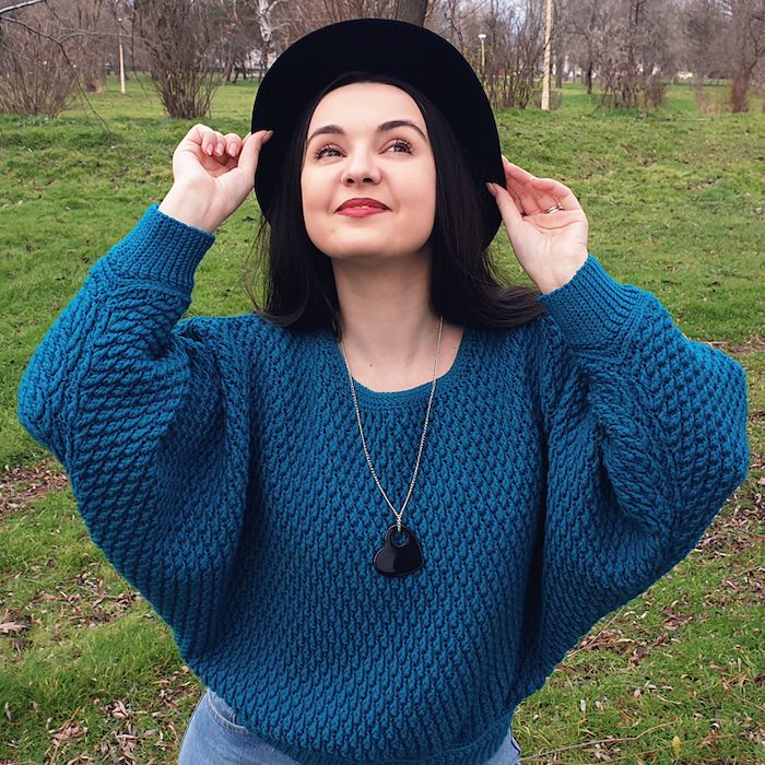A woman in a textured blue crocheted sweater stands with her hands on a wide-brimmed black hat, looking up and to the right and smiling. 