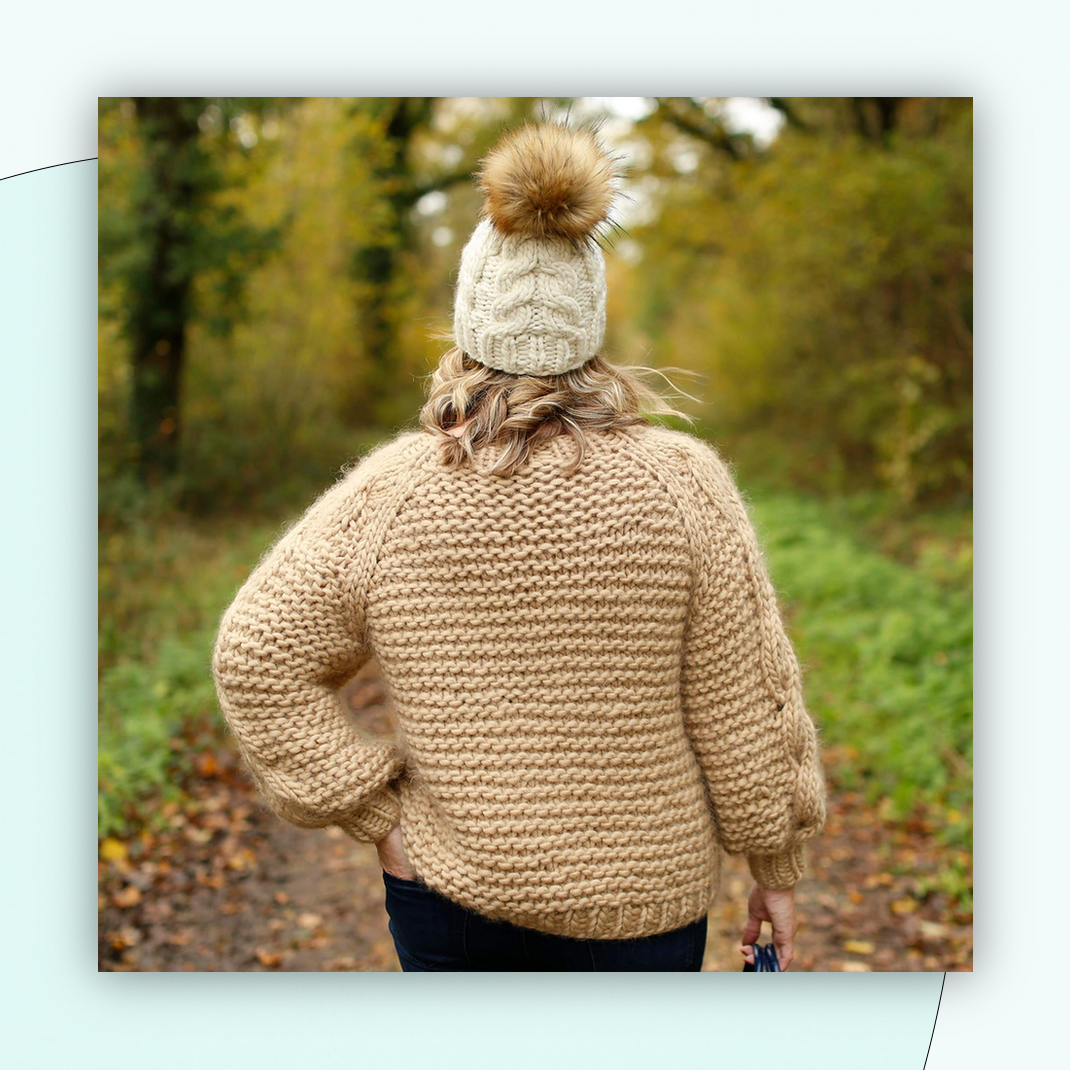 A woman stands on a path in the woods, with her back to the camera, looking down the path, her left hand in the back pocket of her dark jeans. She is wearing a handknit bulky sweater in a light peachy tan color with voluminous sleeves which have a cable running up the length of them. Her curly blonde hair peeks out of a cream cable-knit hat with a puffy fur pom-pom.