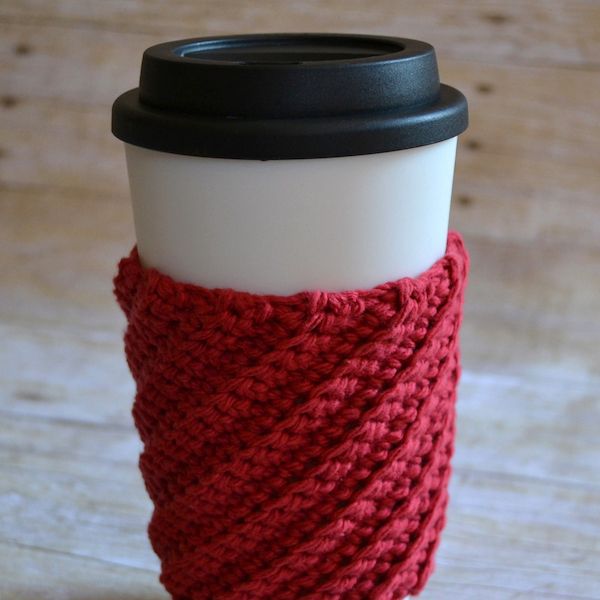 A takeaway coffee cup is on a light wooden table. It has a black lid and a red diagonally textured crocheted cover.