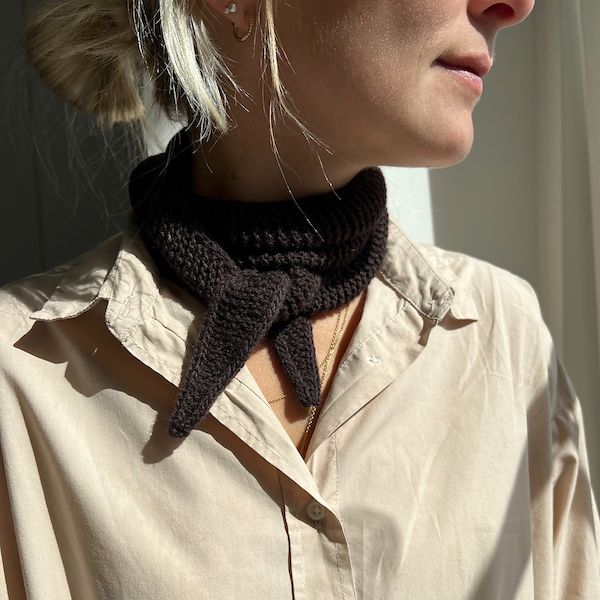 A woman with blonde hair looks offscreen. She is wearing a light tan button down shirt and a knotted handknit scarf.