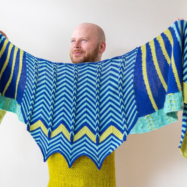 Knitwear designer Stephen West holds up a boldly striped shawl in shades of royal blue, aqua, and chartreuse.