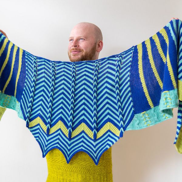 Knitwear designer Stephen West holds up a boldly striped shawl successful shades of royal blue, aqua, and chartreuse.