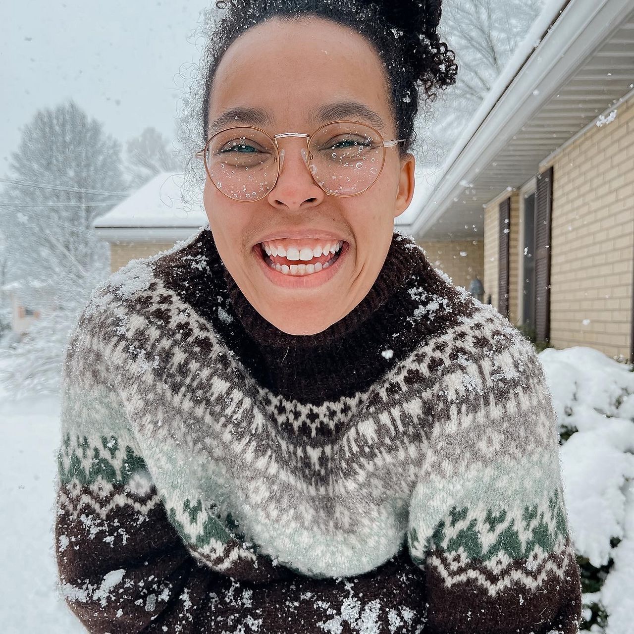 a woman stands in the snow, grinning at the camera, with snowflakes falling against her clothes, hair, and glasses. her sweater is stranded colorwork knitting with a yoke in shades of black, white, charcoal, light grey, mint, and dark aqua.