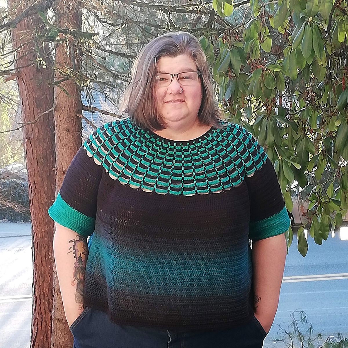 A woman stands in front of a tree, looking at the camera confidently with a small smile. Her crocheted sweater has gradient colors of black and teal on the bottom, and the yoke is worked in an open textured pattern with stripes of black and turquoise.