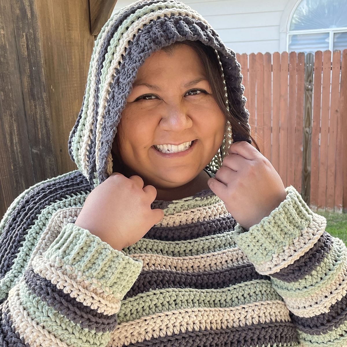 a woman smiles broadly at the camera. she wears a striped crochet hoodie sweater in charcoal, cream, and mint screen.