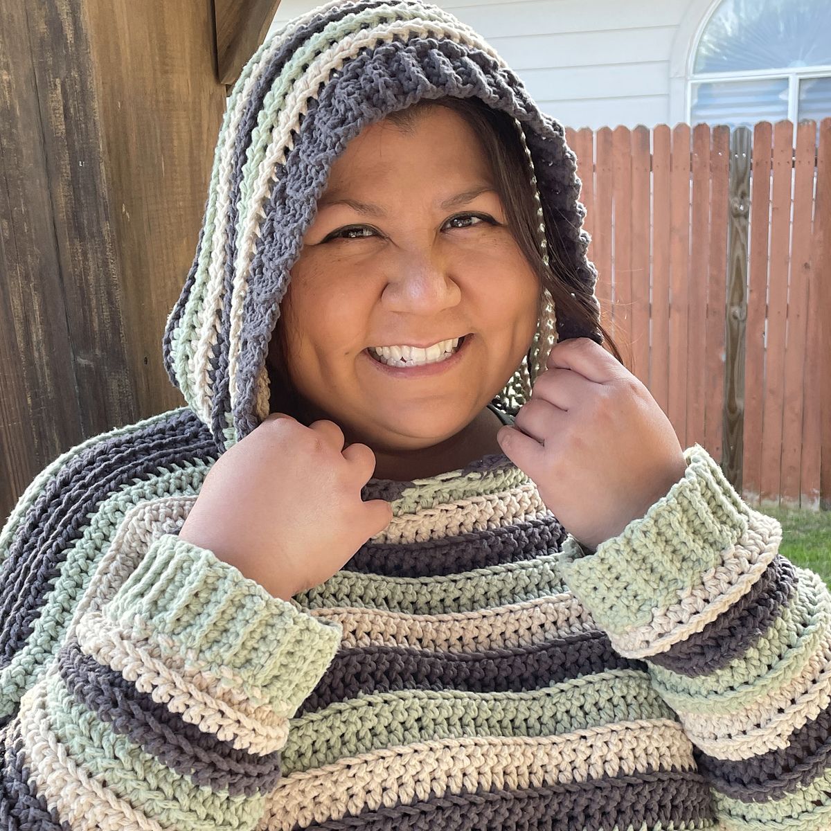 a female smiles broadly astatine nan camera. she wears a striped crochet hoodie sweater successful charcoal, cream, and mint screen.