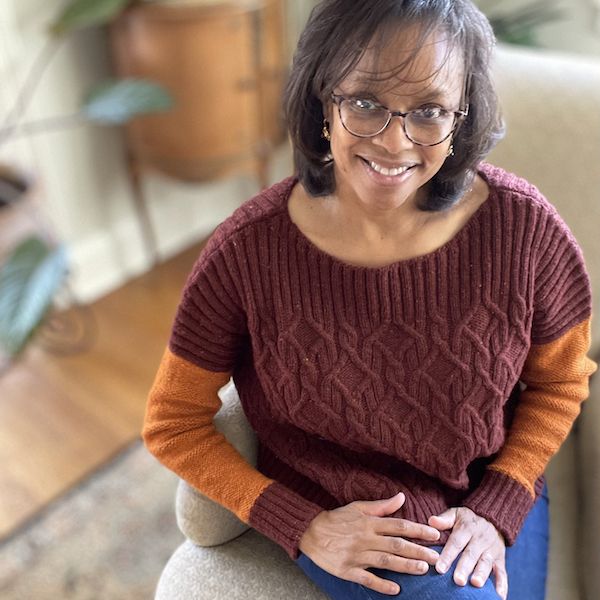 a woman sits on a chair, smiling and looking up at the camera. she wears a cabled sweater in burgundy with orange sleeves.