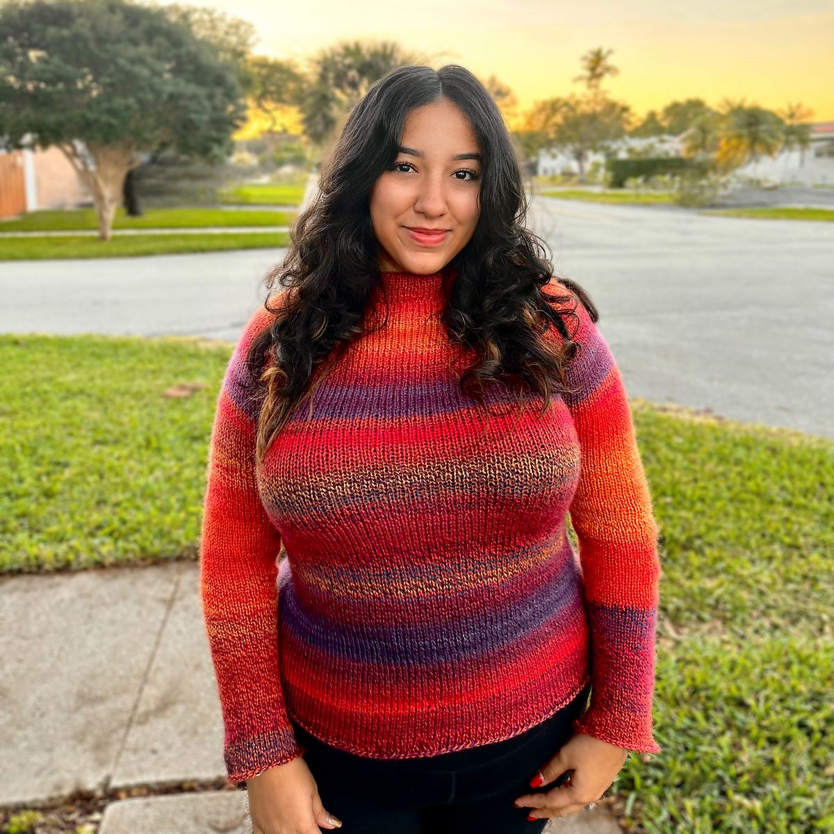 a woman stands directly in front of the camera at sunset. she wears a sweater in striped yarn with shades of red, pink, orange, and purple, which glows in the golden hour light.
