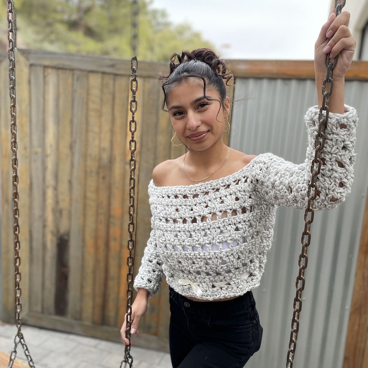 a woman wearing a lace crocheted sweater in a chunky white yarn stands with her hands on swing chains, smiling directly at the camera