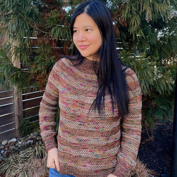 a woman stands in front of an evergreen tree, smiling and looking to the distance. she wears a variegated sweater with wide bands of stockinette alternating with textured bands