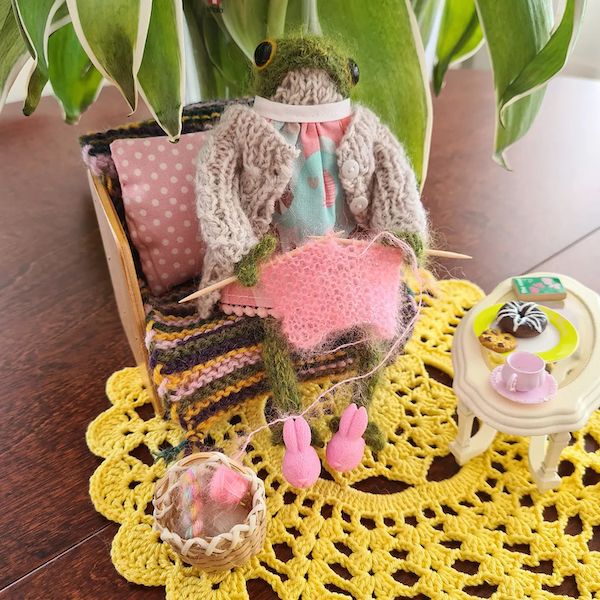 A little knit frog sits on a tiny chair, dressed in a dress and knit sweater, and posed with tiny knitting in its hands. A little table with doll food - cookies, donuts, and tea - is in front of the frog.