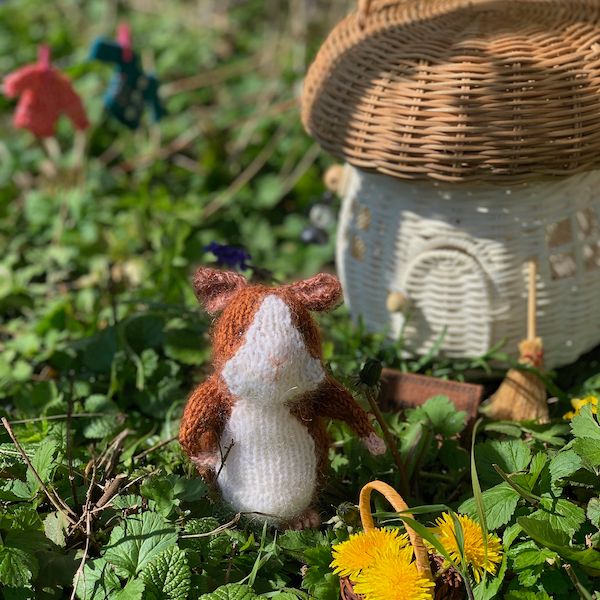 a tiny handknit guinea pig in front of a wicker mushroom-shaped house with a little broom propped on it. In the background is a laundry line with little sweaters.