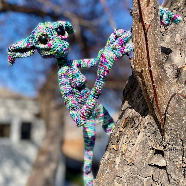 A crocheted multi-colored sprite with long legs, large eyes, and pointed ears, peers around the branch of a tree.