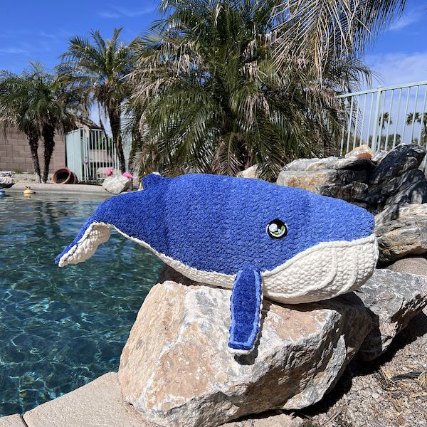 A large crocheted humpback whale stuffie rests on top of a rock by a shimmering blue pool, palm trees in the background.