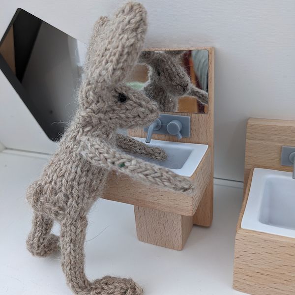 A tiny knit bunny leans over a little toy sink, seeming to wash its hands while it looks at its reflection in the toy mirror.