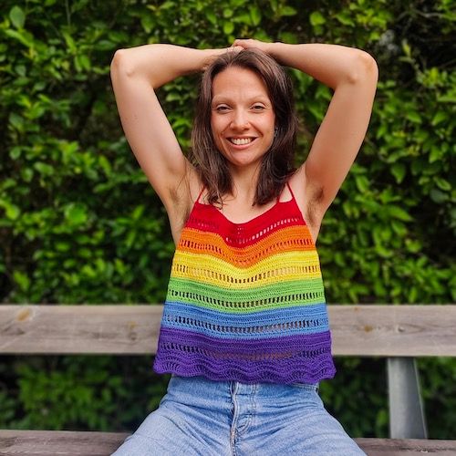 A woman sits on a bench in front of greenery, hands behind her head, smiling confidently at the camera. She is wearing a crocheted camisole worked in bright rainbow stripes.