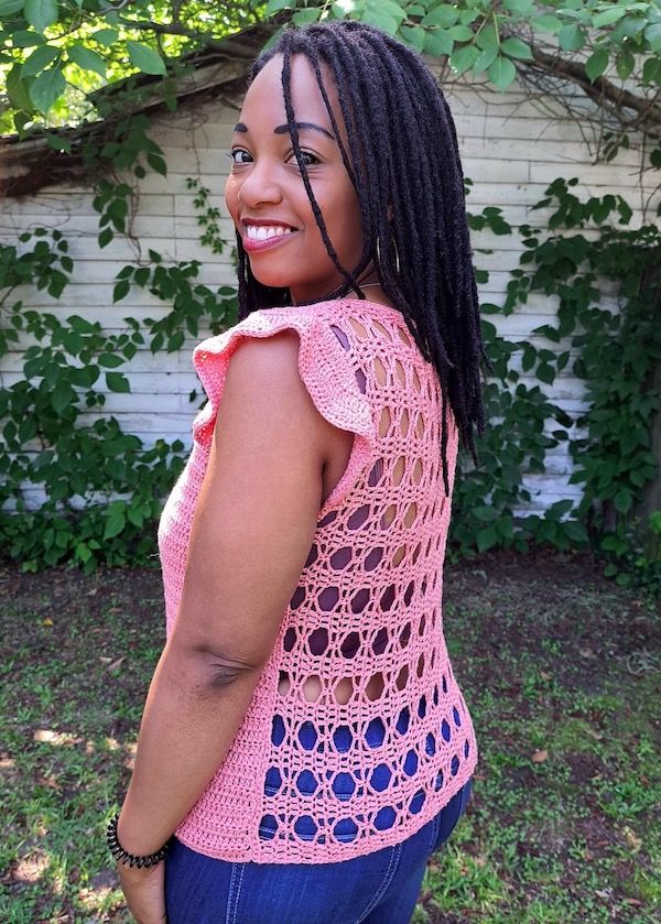 A woman looks over her shoulder with a sunny smile, showing off the openwork back of her bright pink crocheted top.