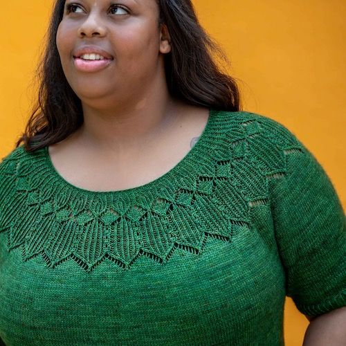 A smiling woman looks up, posed against an orange wall in a deep green handknit sweater with a lacy circular yoke.