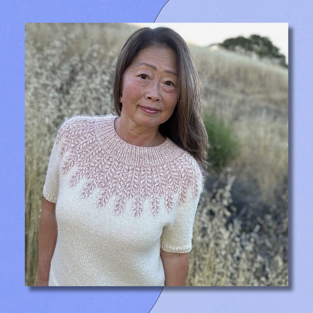 A woman stands on a golden, grassy hill, greying hair flowing and a warm expression on her face. She is wearing a beautiful handmade cream sweater with a mauve colorwork yoke.