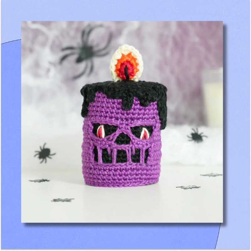 A crocheted purple skull candle, complete with crocheted flame, on a white background with black spiders.