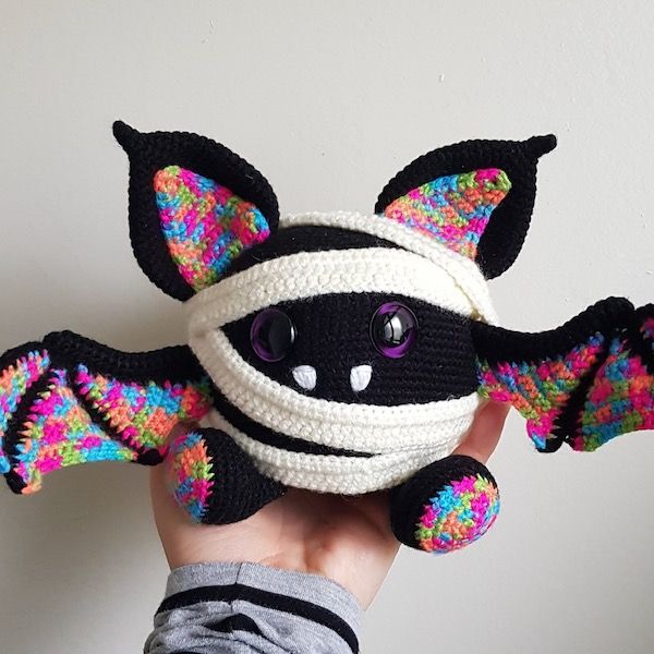A hand holds up a pleasingly round crocheted bat in a mummy costume. It has purple eyes and colorful variegated yarn for the insides of its ears and wings, and the bottom of its feet.