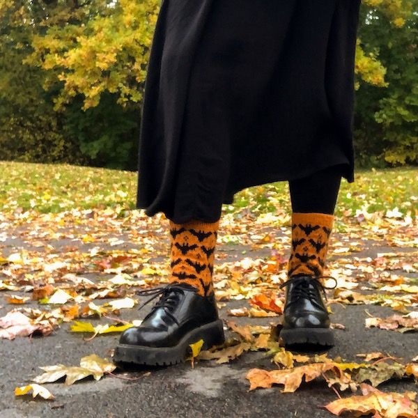 A person's lower half, clad in a long black skirt, black tights, and chunky black shoes, showcasing orange knit socks with black bats worked in stranded colorwork. Behind them is an open field with fall leaves on the ground and fall colors in the trees in the background.
