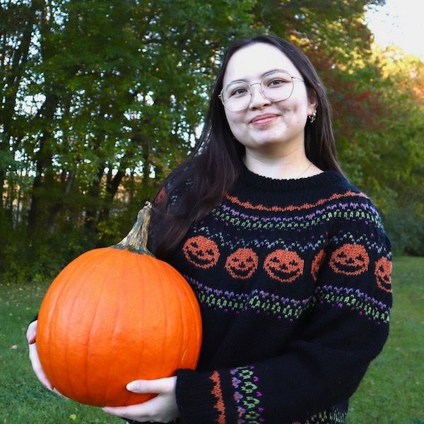 A person smiles at the camera, holding a pumpkin and wearing a stranded colorwork knit sweater with jack o lanterns as the featured design in the round yoke.