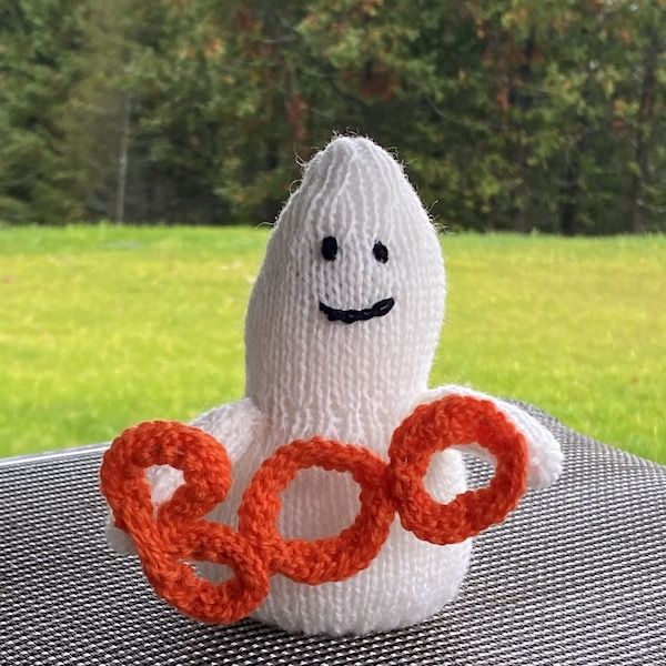 A smiling knit ghost softie holding orange icord letters that spell out BOO!