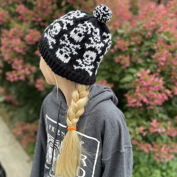 A girl with a long blonde braid looks away from the camera, wearing a knit colorwork hat in chunky black yarn with white skulls and crossbones.