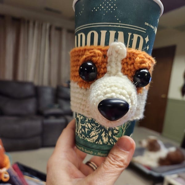 A hand holding a coffee cup with a cozy cup holder that looks like an earnest corgi's face.
