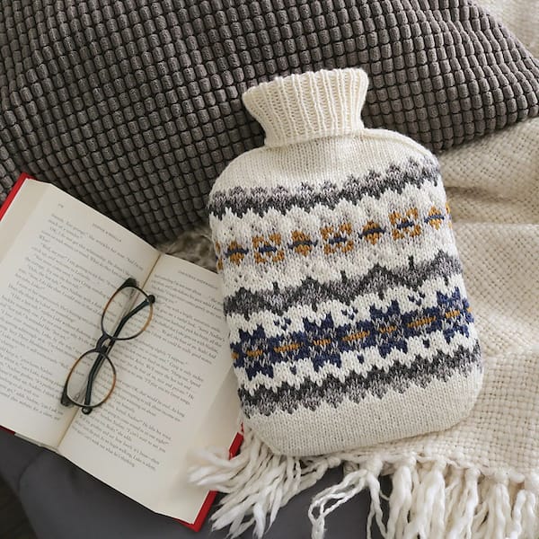 A stranded colorwork hot water bottle cozy in neutral tones likes on a flat-lay with a book, glasses, and textured grey blankets in cream and grey.