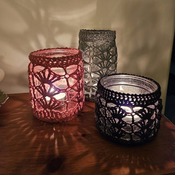 Three mason jars in varied sizes, with tea lights glowing inside. The mason jars are covered in delicate crocheted cozies in red, grey, and black.