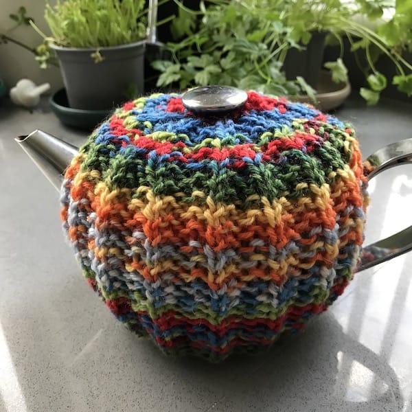 A silver teapot with a mistake rib cozy knit in variegated yarns in colors of red, green, orange, yellow, and blue.