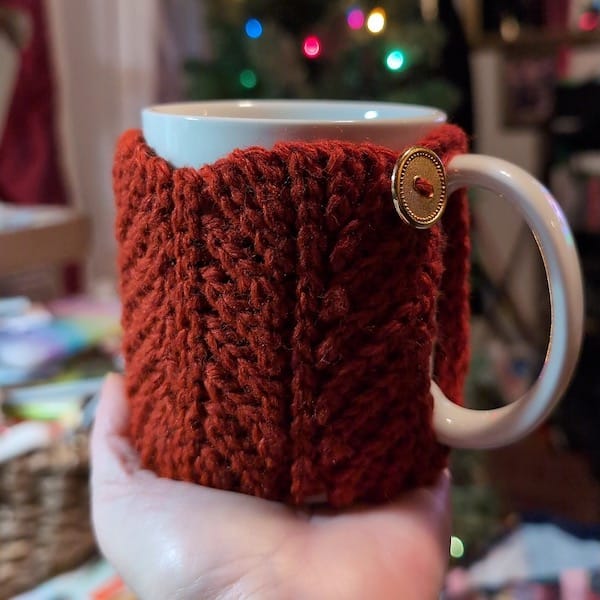 An outstretched hand holding a white mug with a rust-colored textured knit cozy on it, held closed with a brass button.