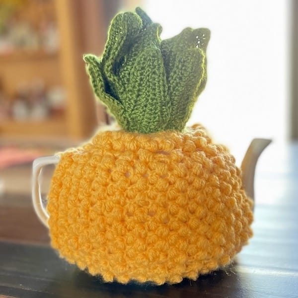 A white teapot covered in a crocheted cozy made to look like a pineapple.