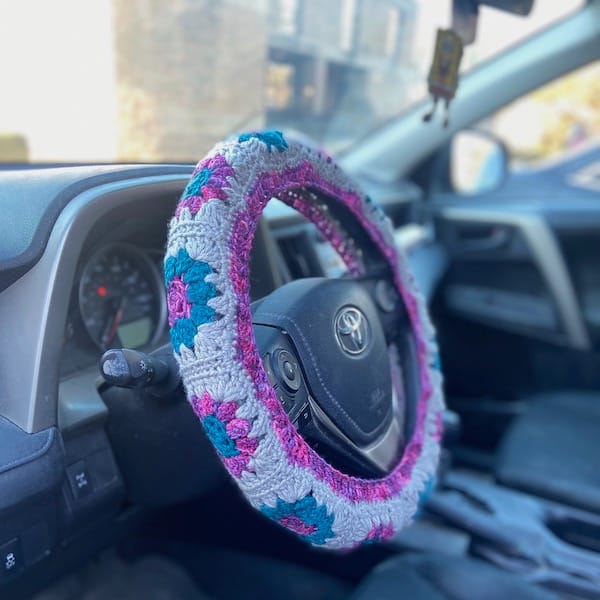 A car steering wheel covered in a granny square cozy in cream, bright pink, and turquoise.