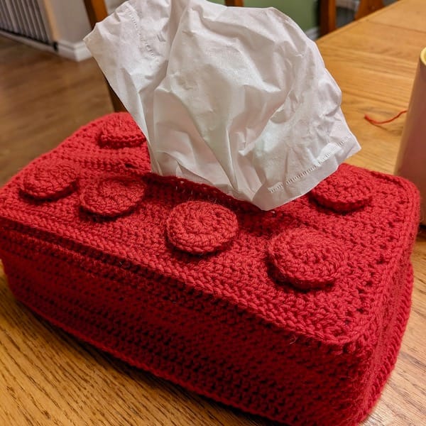 A tissue box with a crocheted cozy resembling a rectangular red lego brick.