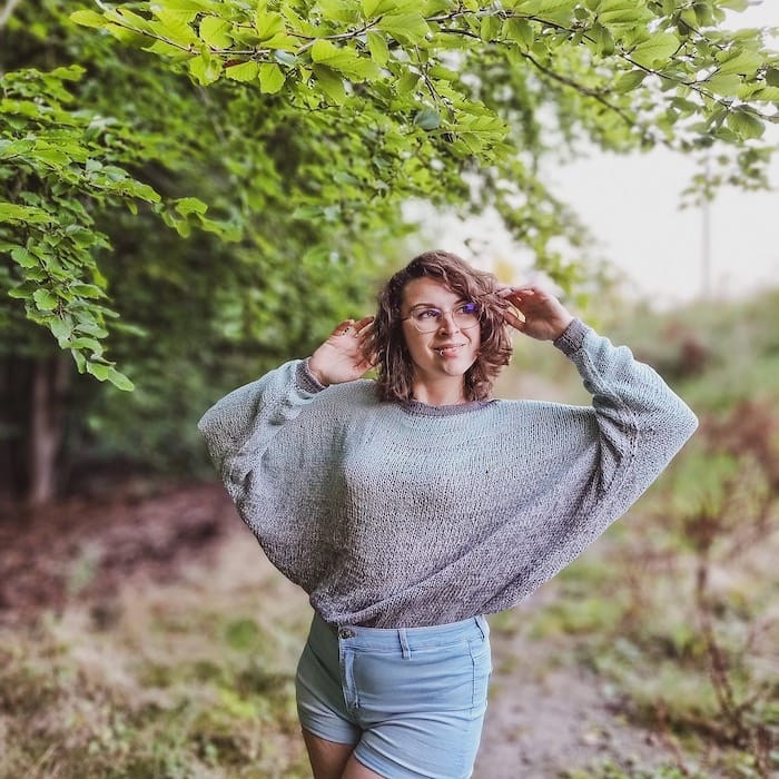 A woman holds up her arms along a wooded path, showcasing the dramatic batwing sleeves of her sweater.