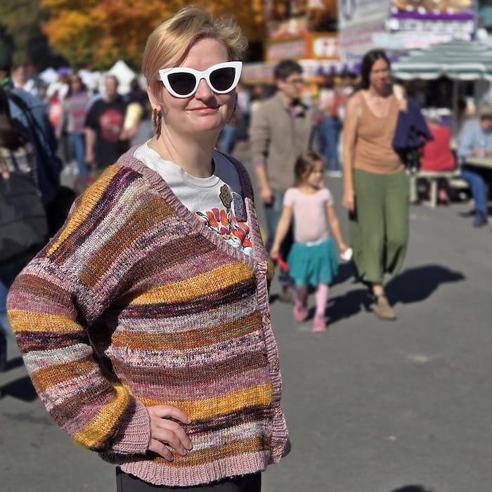 A woman in sunglasses stands smiling softly at the camera, wearing a striped cardigan in shades of browns, pinks, and oranges.