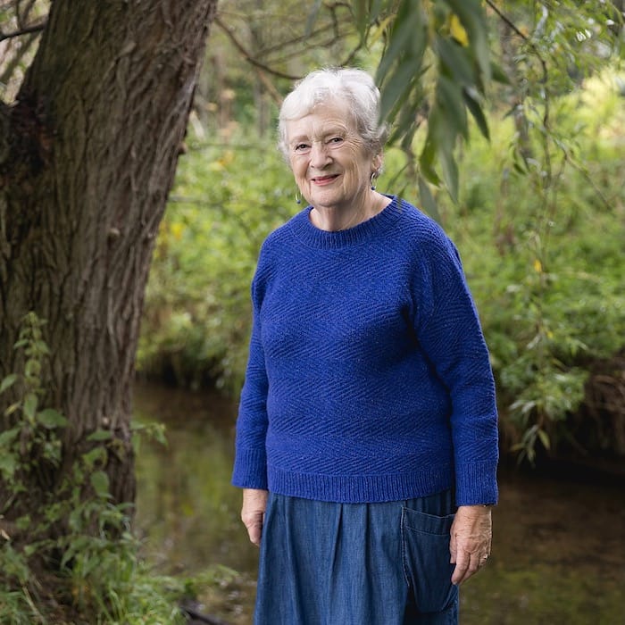 A woman confidently smiles, standing near a tree by a creek, wearing a classic pullover sweater in a bold sapphire blue.